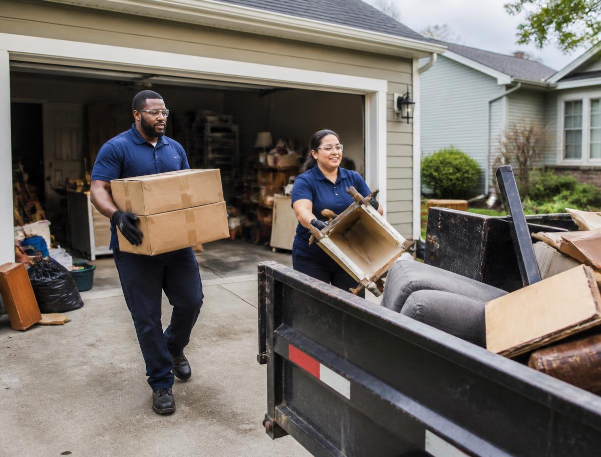 Two uniformed crew members carrying boxes and old furniture out of a home garage to a dump trailer
